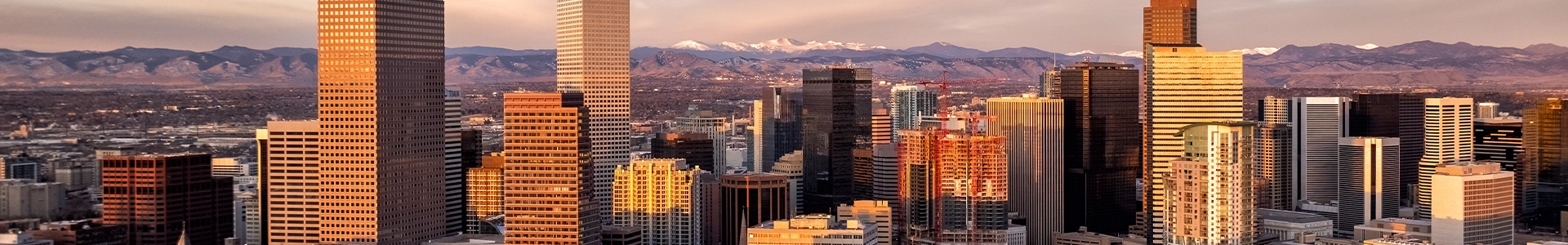 Panoramic view of downtown Denver at sunset showcasing skyscrapers with the Rocky Mountains in the background.